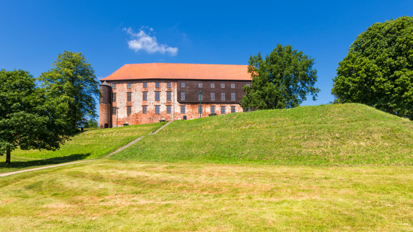 Koldinghus in Kolding mit rotem Ziegeldach, rundem Turm und Fassade aus Backstein und dunklem Holz auf einem grünen Hügel unter blauem Himmel