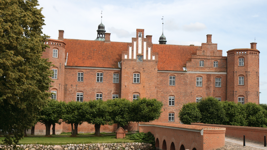 Gammel Estrup auf Djursland mit roter Backsteinfassade, Treppengiebel mit Uhr, runden Ecktürmen und Spitzen, gestutzten Bäumen und Backsteinbrücke im Vordergrund